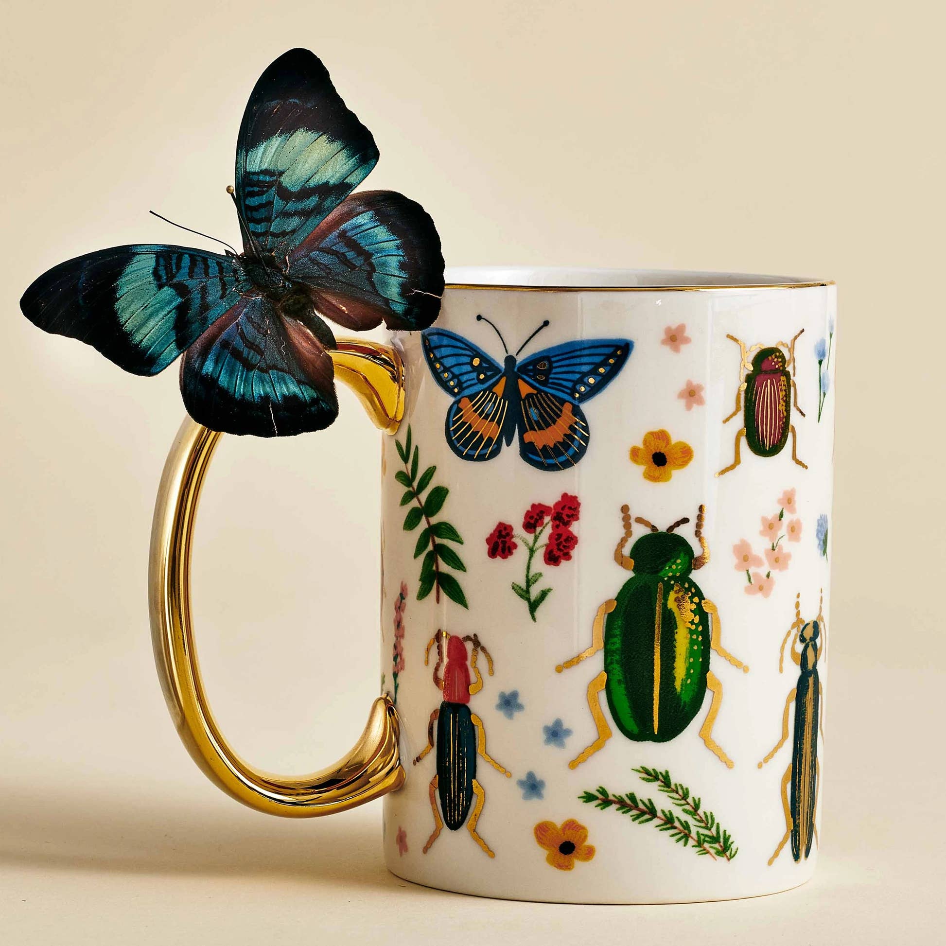 Rifle Paper Co Curio Porcelain Mug illustrated with flowers, insects, and gold accents. A blue butterfly is resting on the gold handle.