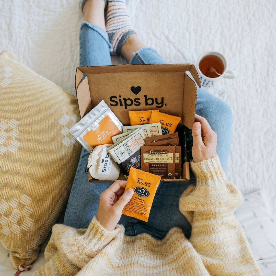 Overview of a person wearing an orange sweater, holding a sample of tea with an open Sips by Box in their lap and a full cup of tea by their side