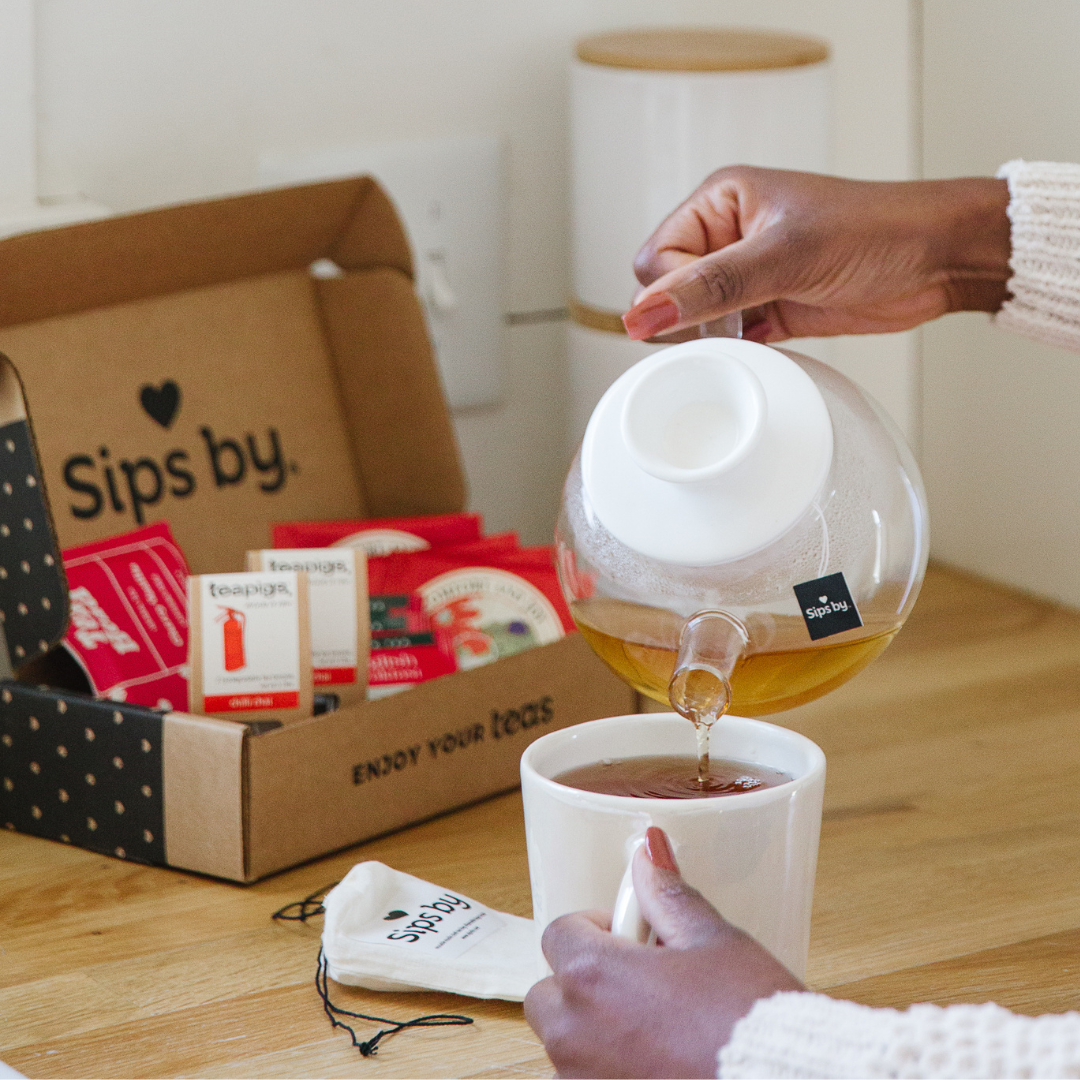 Close up of a person pouring tea from a glass teapot into a mug in a kitchen with an open Sips by Box on the counter