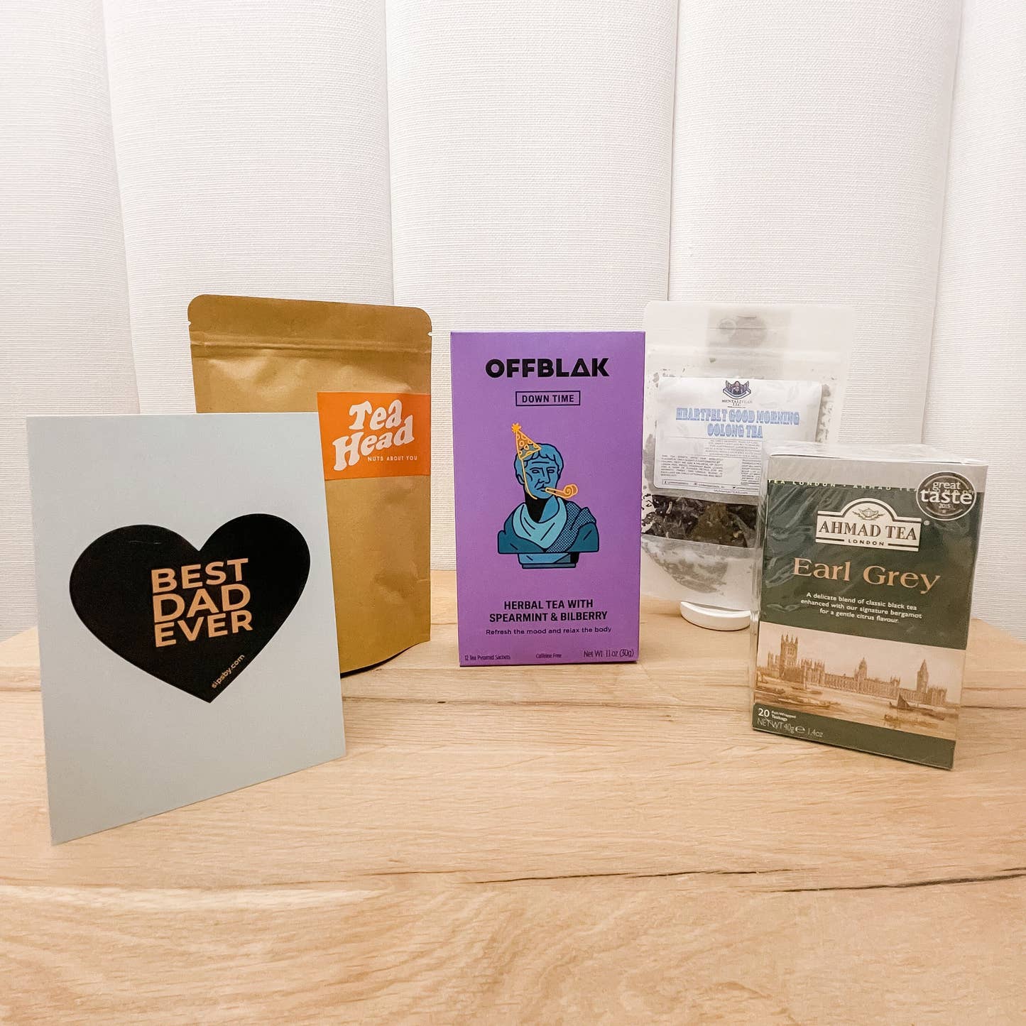 Four full-sized tea packages from OFFBLAK, Tea Head, Mentaliteas, and Ahmad Tea for the Father's Day Tea Care Package and a Sips by "Best Dad Ever" blue postcard with a heart