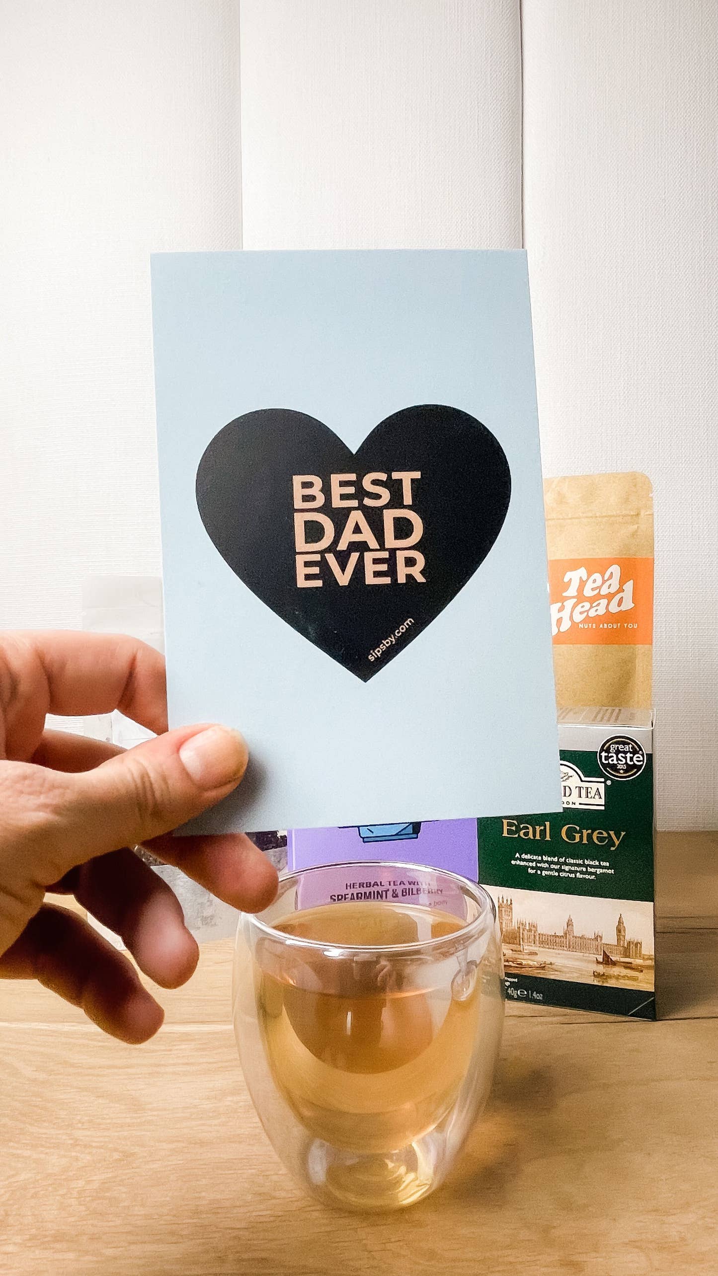 Hand holding the Sips by "Best Dad Ever" blue postcard with a black heart in front of Father's Day Care Package teas and a brewed cup of tea in the iridescent glass mug
