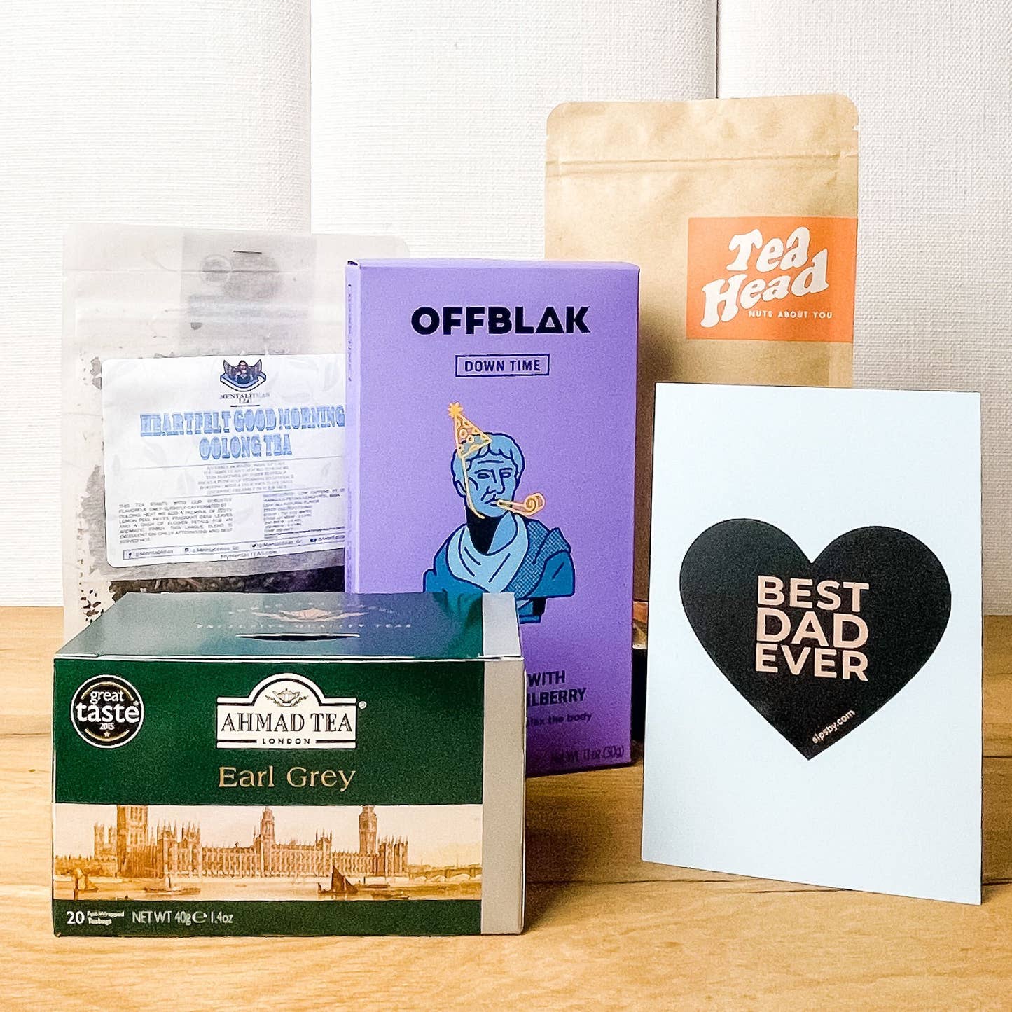 Four full-sized tea packages from OFFBLAK, Tea Head, Mentaliteas, and Ahmad Tea for the Father's Day Tea Care Package and a Sips by "Best Dad Ever" blue postcard with a heart
