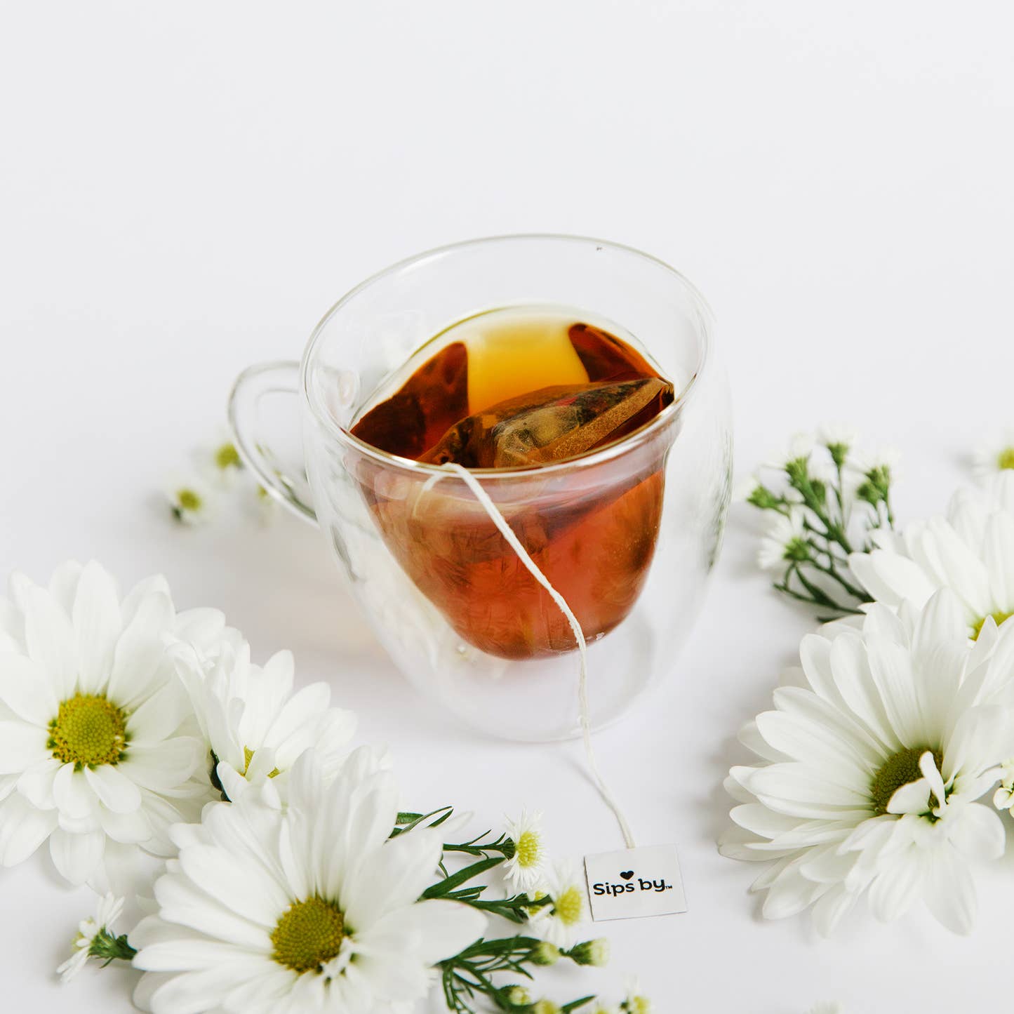 Double-walled glass heart mug with tea from sips by and surrounded by flowers