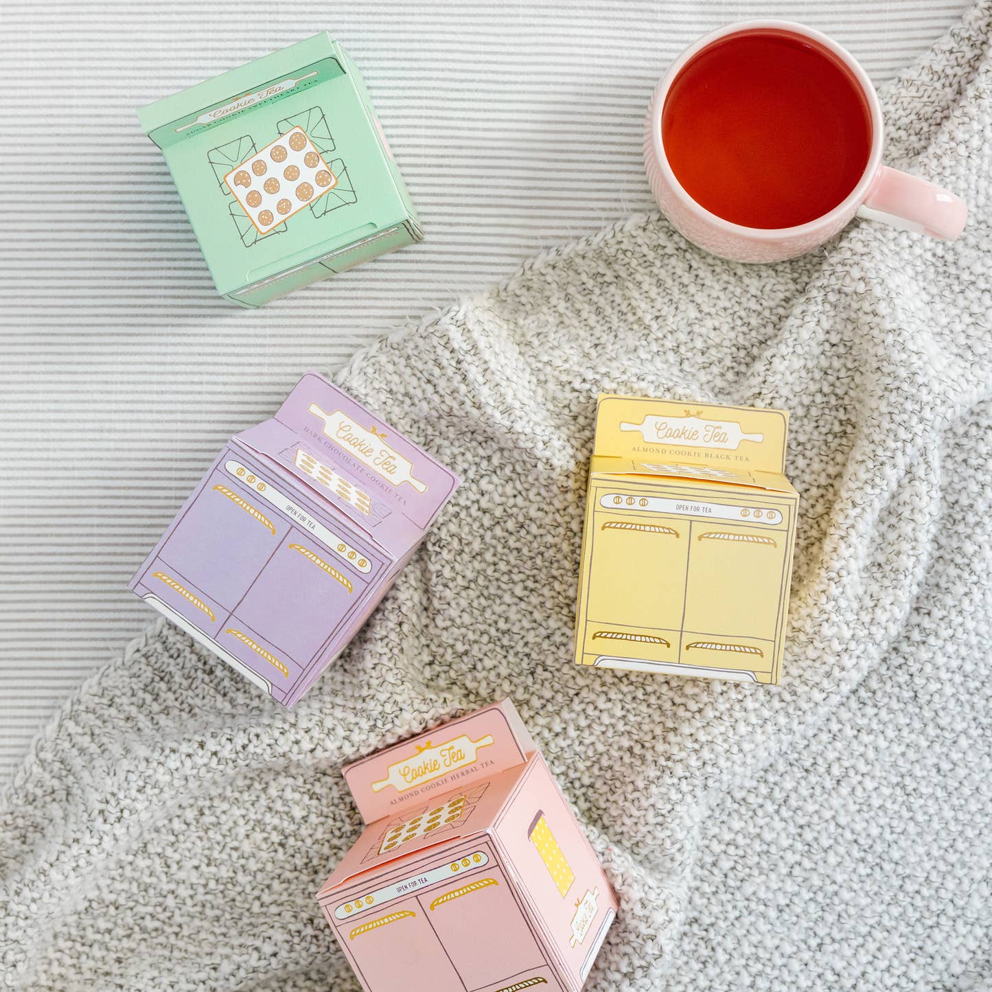 Pink, green, yellow, and purple cookie tea ovens displayed on gray blanket with a cup of brewed cookie tea from Sips by